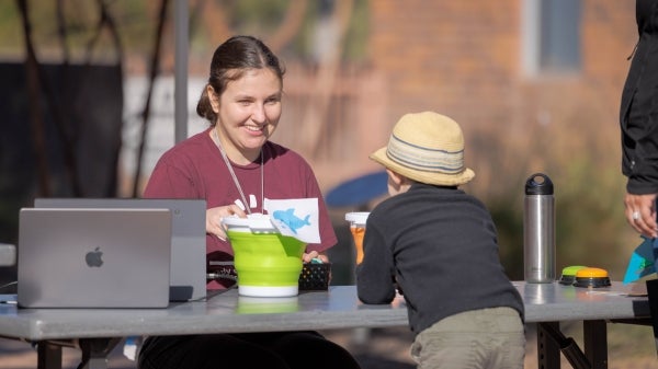 A woman at a table showing a shark illustration to a child wearing a straw hat.