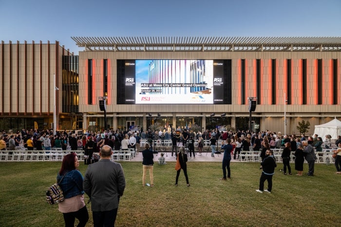 A crow of people sitting and standing in front of a building with a large digital screen